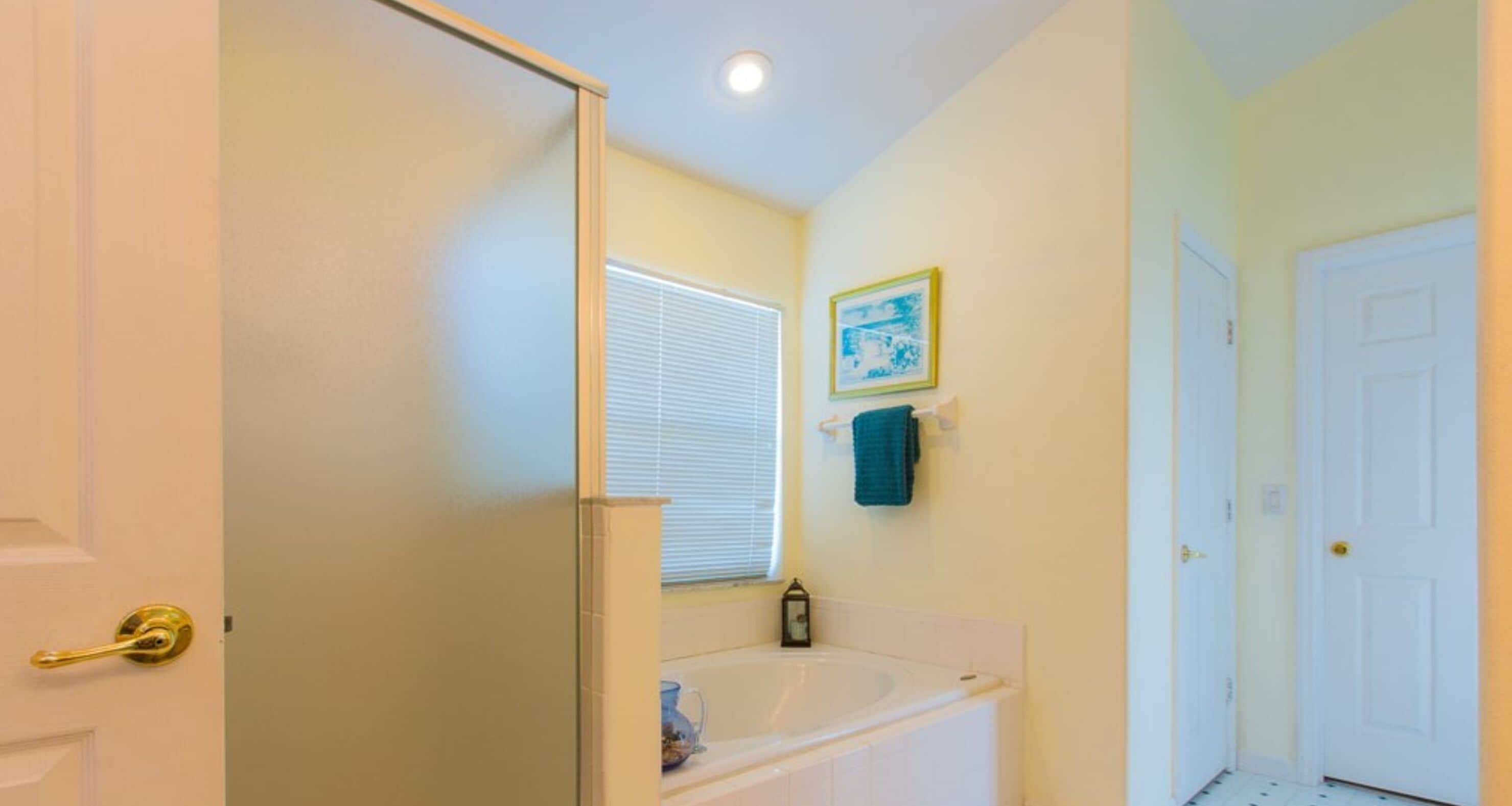 A bathroom with a bathtub, a window with blinds, a towel rack with a towel, and a door.