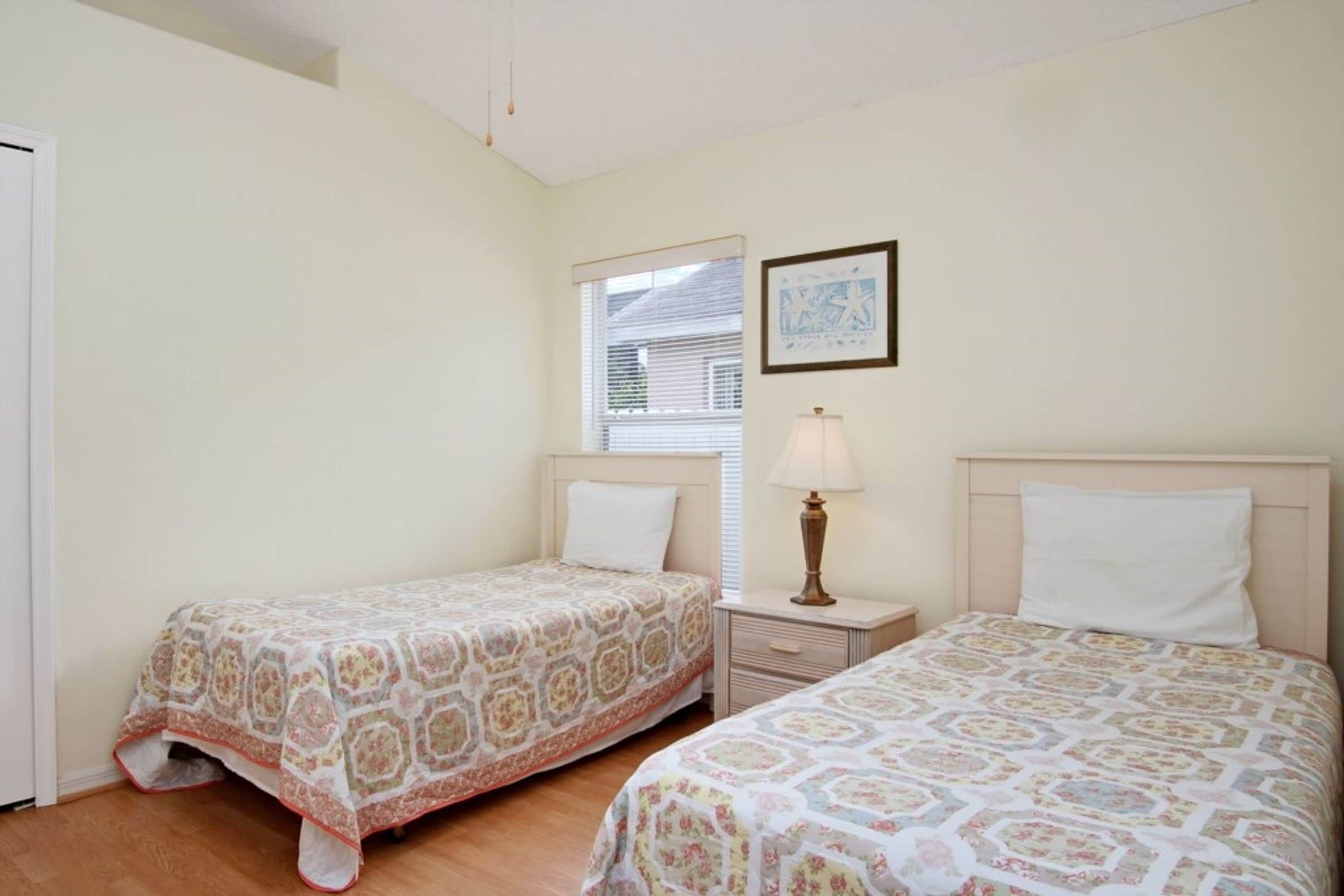 A bedroom with two beds, a nightstand with a lamp, a window with blinds, and a framed picture on the wall.