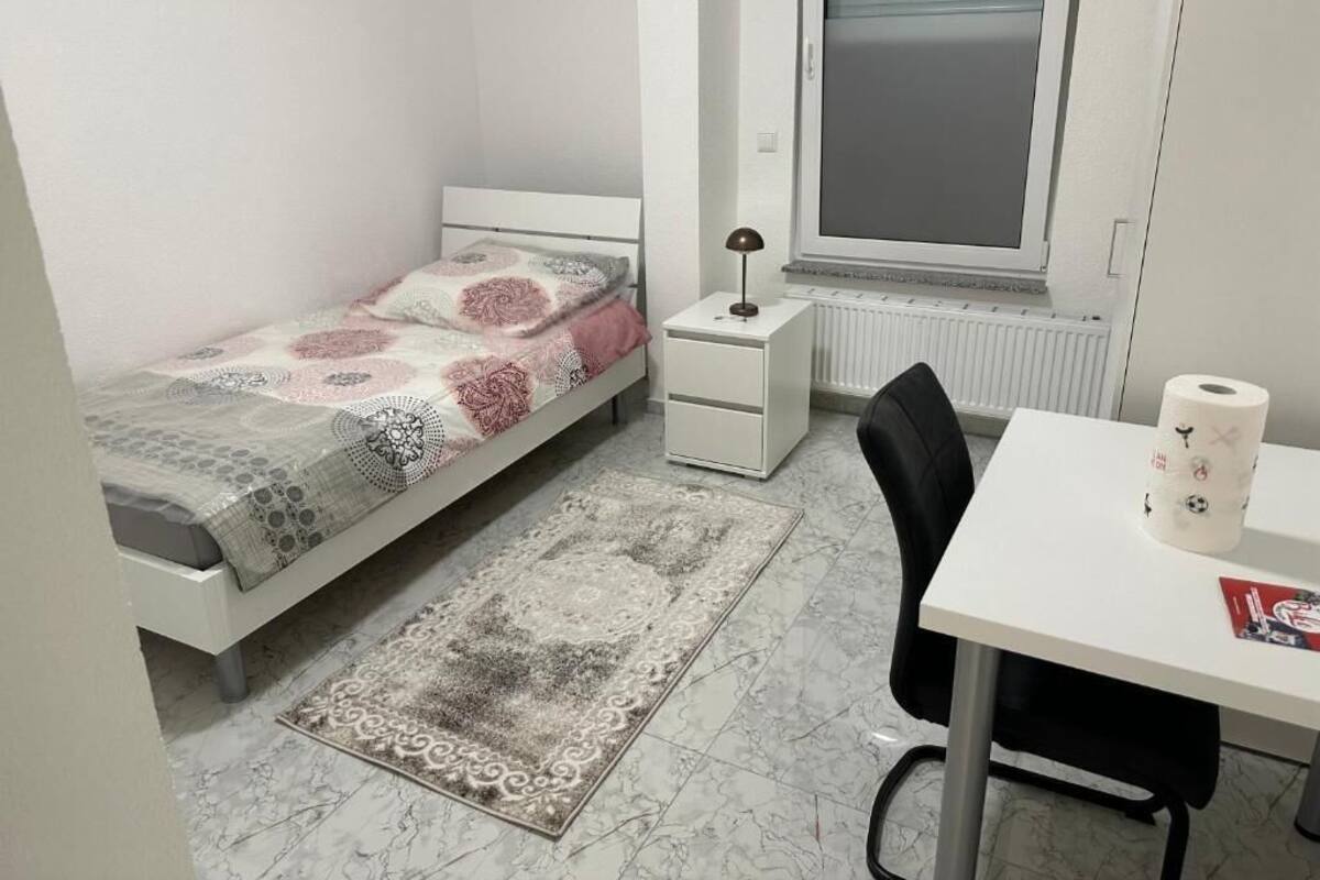 Quarto pequeno e bem organizado, com uma cama de solteiro, uma escrivaninha e uma cadeira.