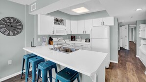 A modern kitchen with white cabinetry, a large island with bar stools, a clock, and a microwave. - Four Winds 304 (Orange Beach)