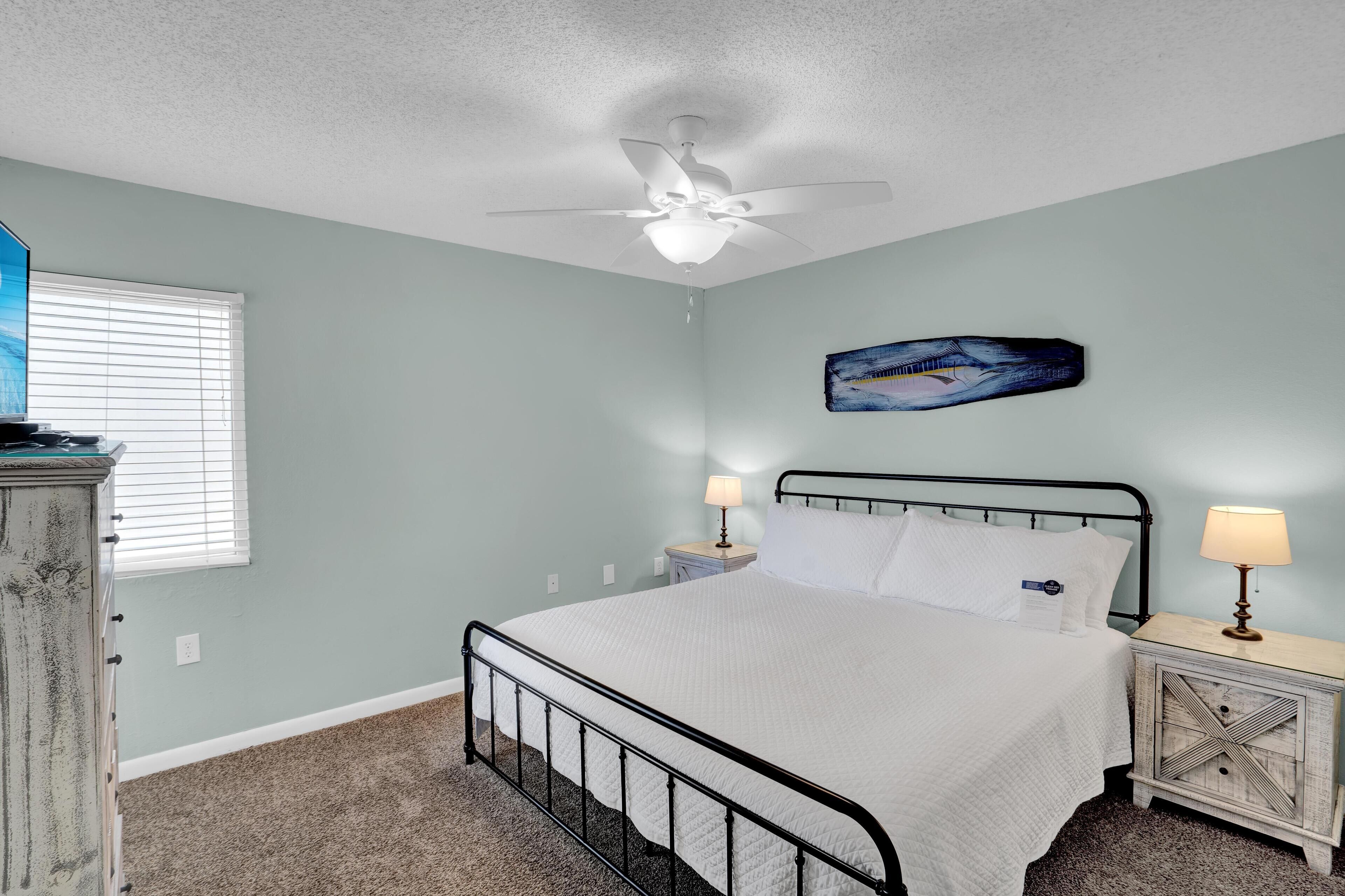 A bedroom with a bed, a ceiling fan, a nightstand with a lamp, a dresser, and a wall-mounted painting.