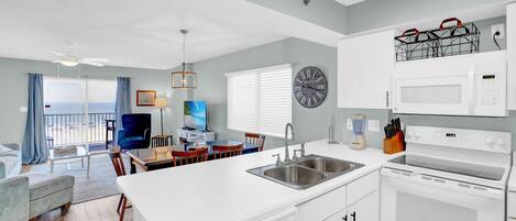 Une cuisine moderne avec des appareils électroménagers blancs, un coin repas avec une chaise bleue et une vue à travers la porte vitrée.