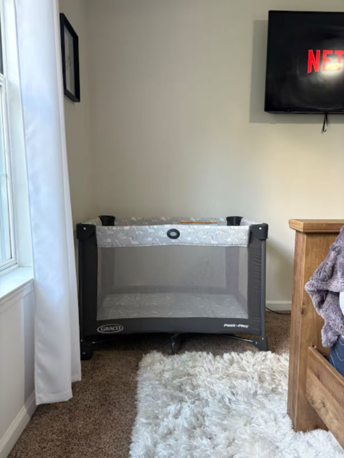 Une pièce comprenant un parc pour enfants de la marque Graco, un tapis blanc et moelleux, une table d’appoint en bois et un téléviseur fixé au mur.
