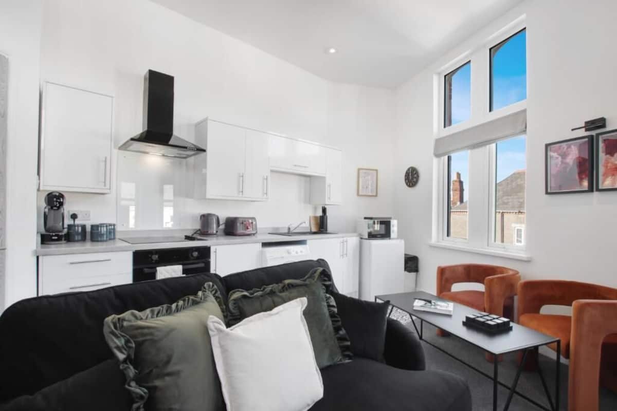 Une cuisine moderne avec des meubles blancs, une hotte noire et un coin repas comprenant une table et des chaises orange.