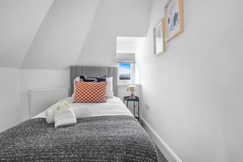 Ein Schlafzimmer mit schrägen Wänden, einem Bett mit grauem Kopfteil, weißer Bettwäsche, einem karierten Kissen und einem kleinen Nachttisch mit Lampe.