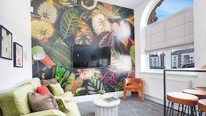 A modern living room with a large TV, a green sofa, a round coffee table, and a vibrant floral mural on the wall.