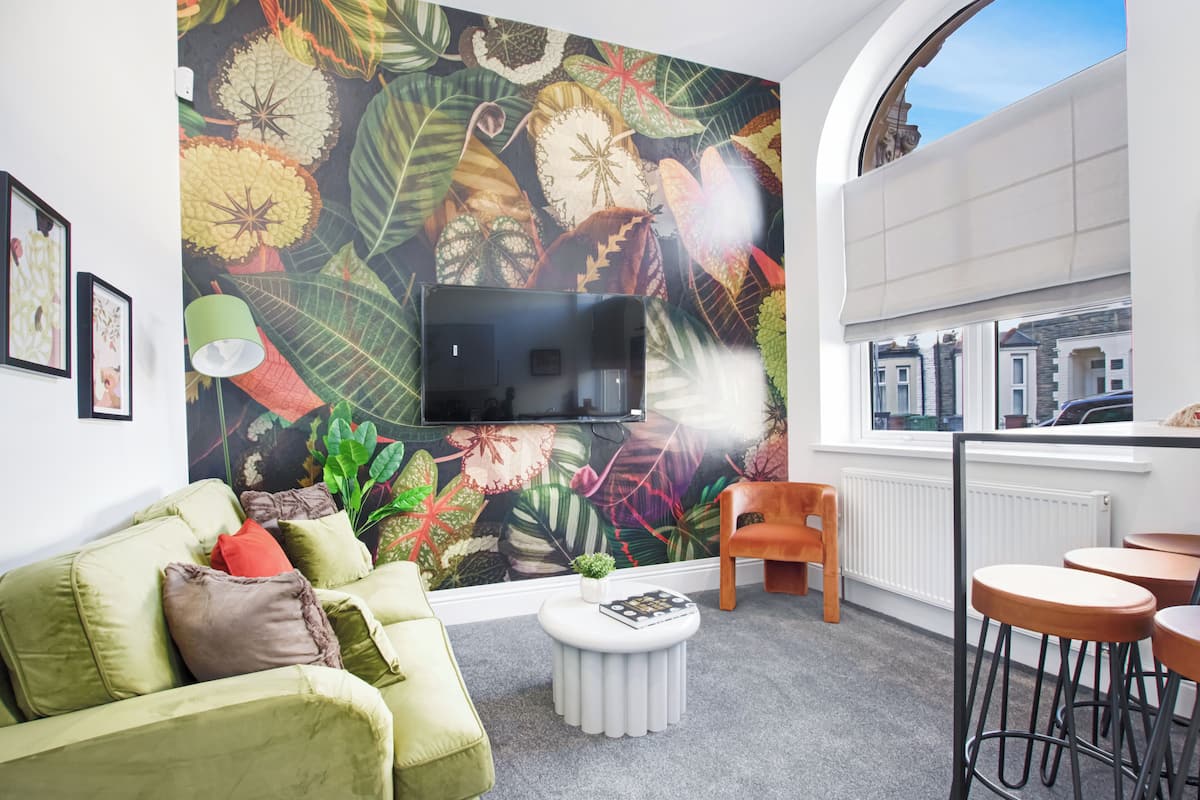Un salon moderne avec un grand téléviseur, un canapé vert, une table basse ronde et une fresque florale aux couleurs vives sur le mur.