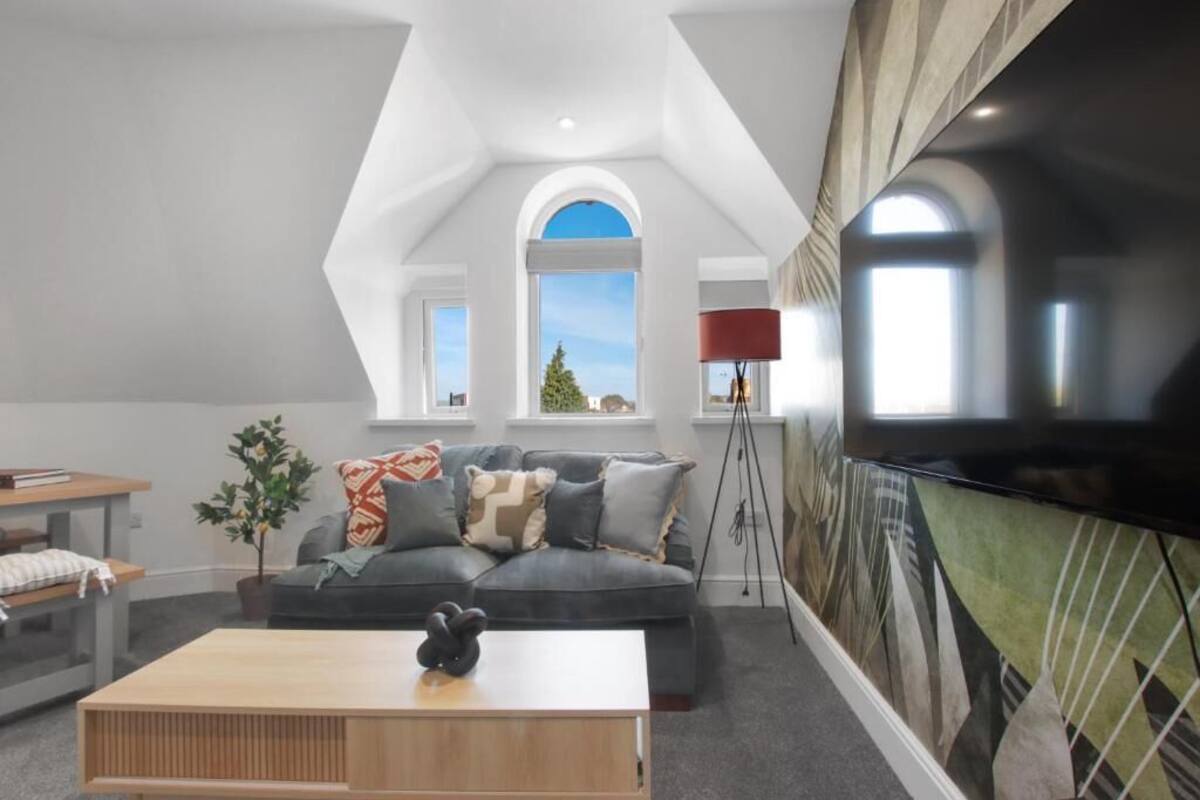 Un salon moderne avec un canapé gris, une table basse en bois et un grand téléviseur à écran plat. On y voit deux fenêtres à arc en plein cintre, et une plante est visible sur la gauche.