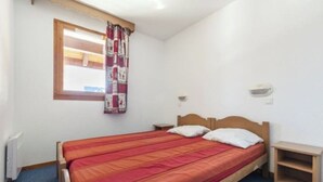 A single-bed room with a wooden headboard, a red bedspread, a window with a curtain, a bedside table, and a wall-mounted light.