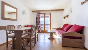 A room with a wooden dining table, chairs, a sofa with red cushions, a small table, and a window with a view of a mountain.