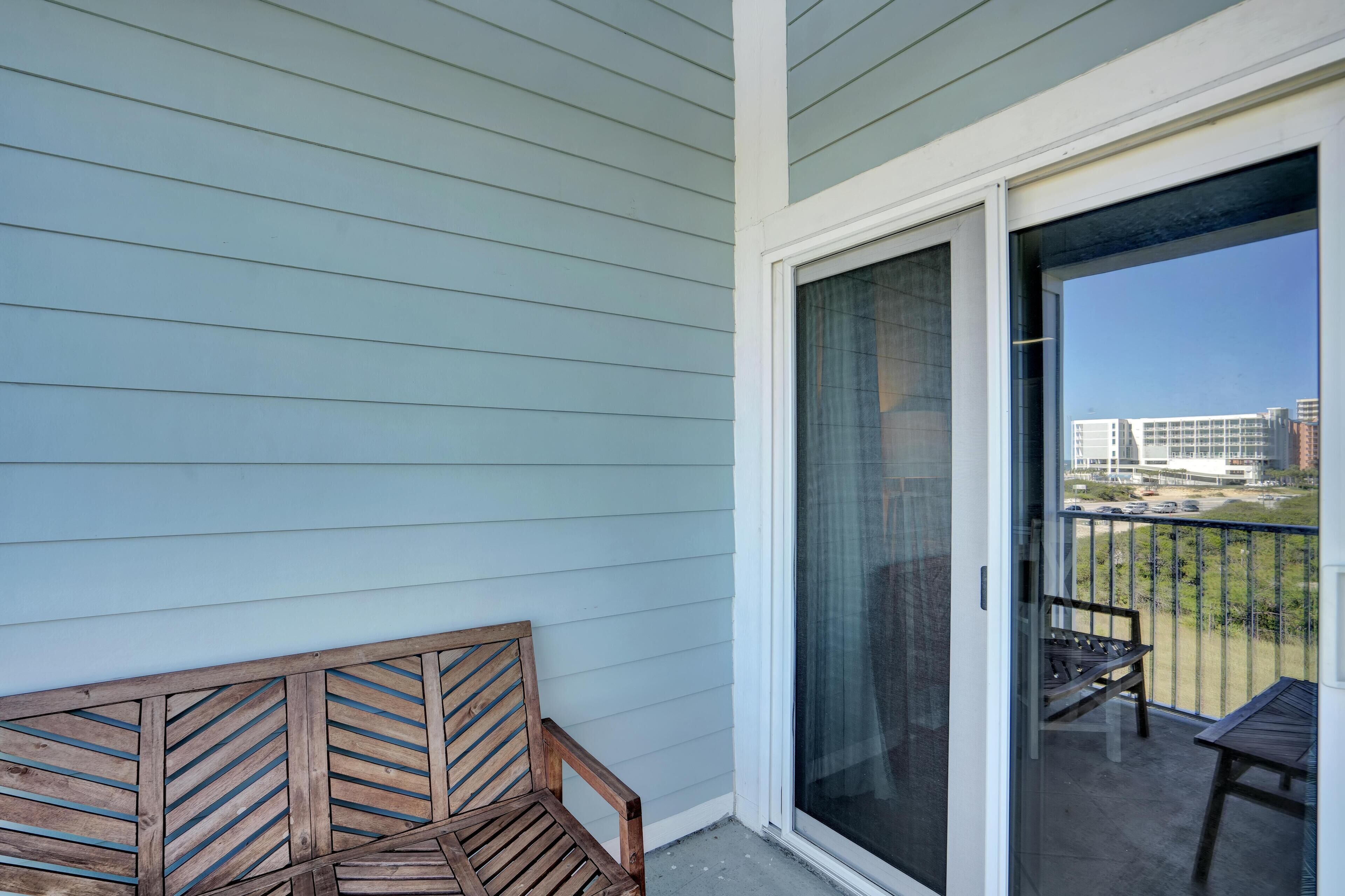 En balkon med træbænk og bord, en skydedør samt udsigt til bygninger og grønt.