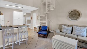 Eine moderne Küche mit weißen Schränken, einer zentralen Kochinsel mit Barhocker, einer Wendeltreppe und einem Wohnbereich mit Sofa und Kissen.