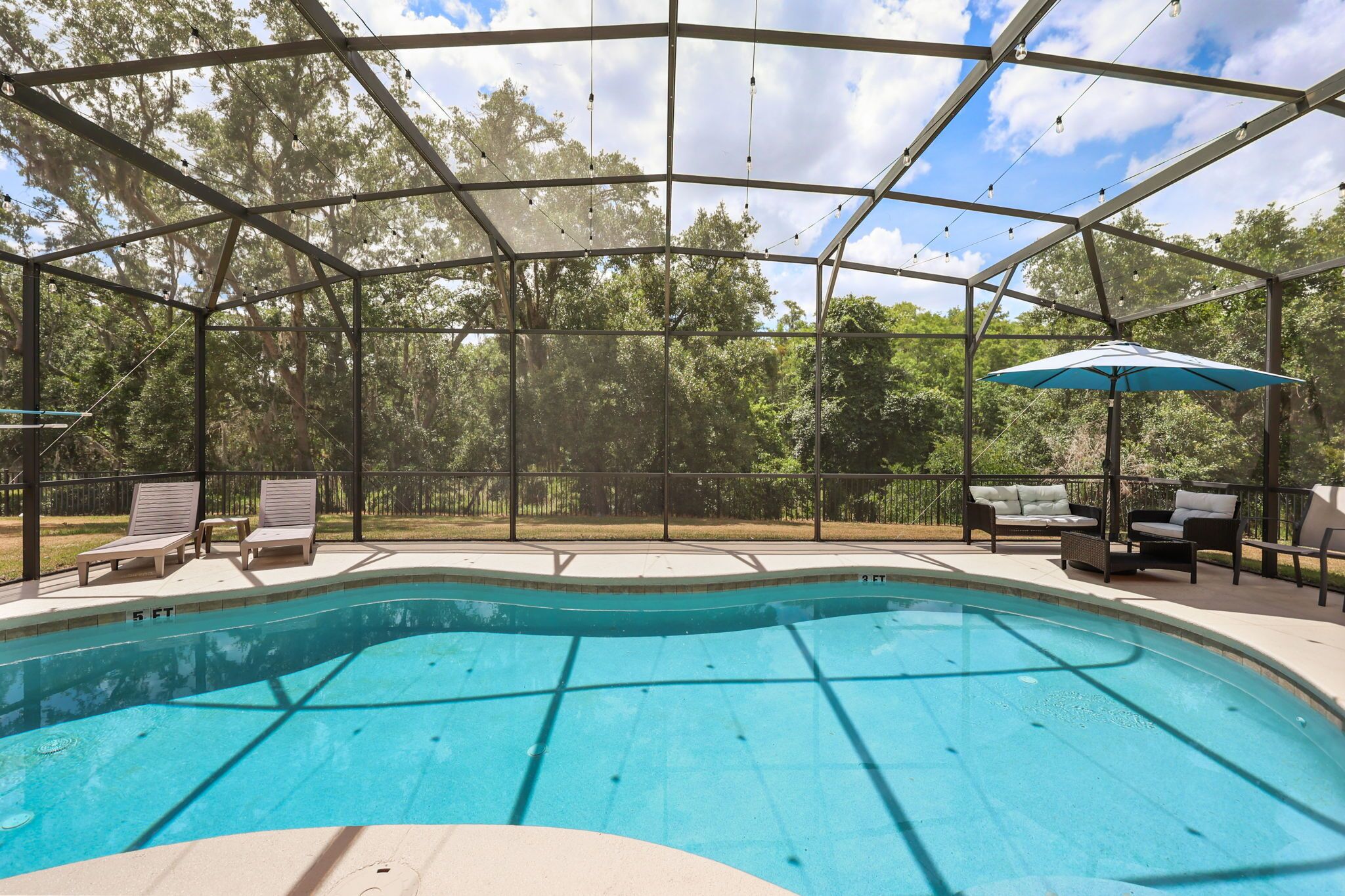 A covered outdoor swimming pool with lounge chairs and a table, surrounded by trees.