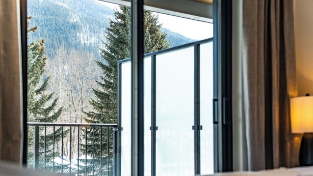 A room with a large window overlooking a forested area and a mountain. - The Peaks and Birdies (Revelstoke)