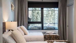 A bedroom with two beds, a window with curtains, a lamp, and a view of trees. - The Peaks and Birdies (Revelstoke)