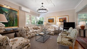 A spacious living room with floral-patterned sofas, a coffee table, and a television. There are decorative lamps, a large painting, and a potted plant.