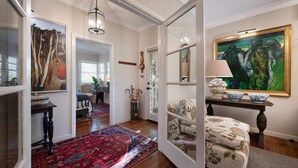 A hallway with a red patterned rug, a wooden floor, a chair, a table with a lamp, and a painting on the wall.