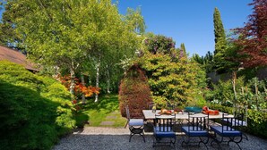 A garden patio with a table and chairs, surrounded by lush greenery and trees.