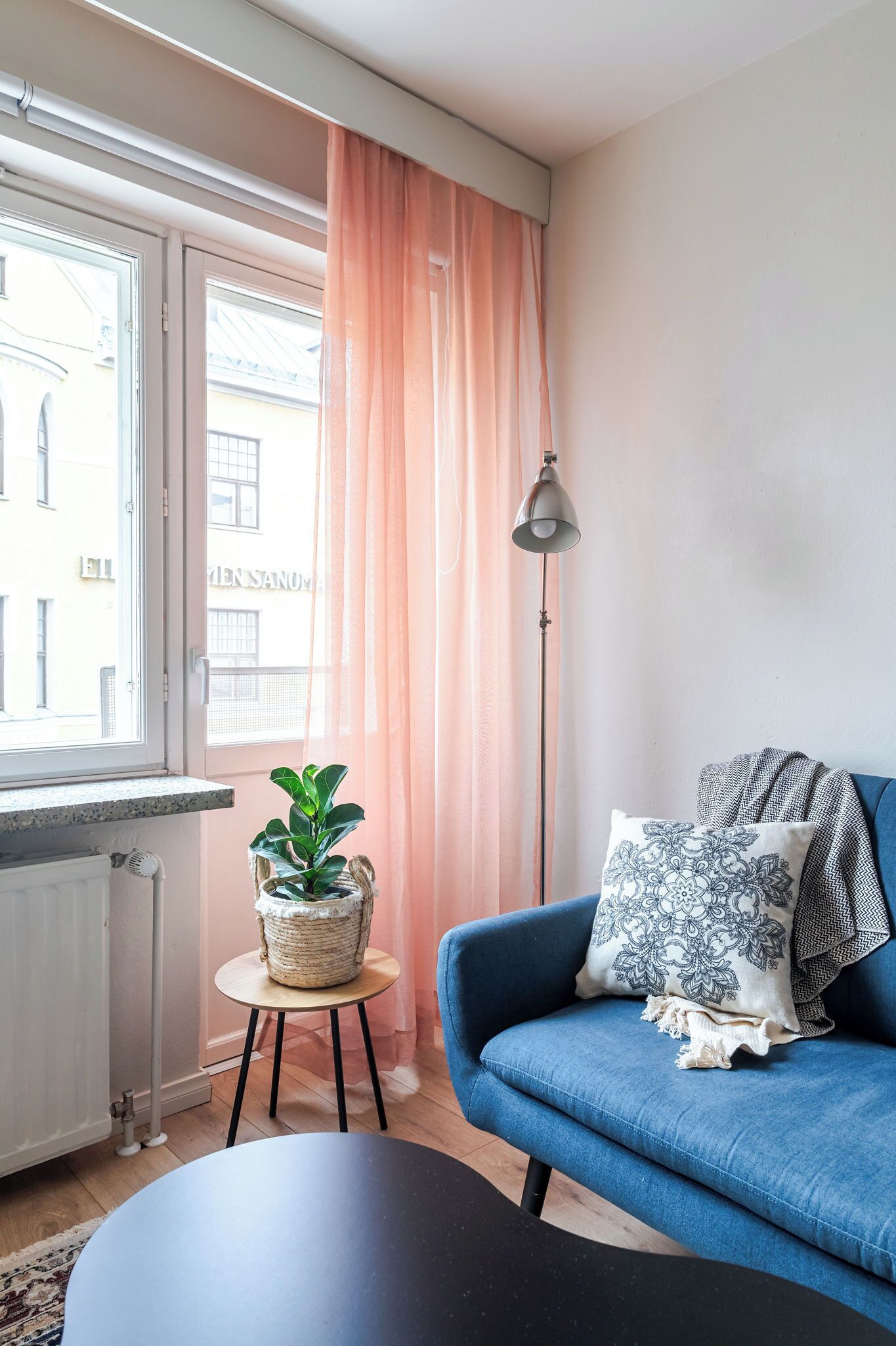A cozy living room with a blue sofa, a small round table with a plant, a floor lamp, and a view of the street through a window with pink curtains.
