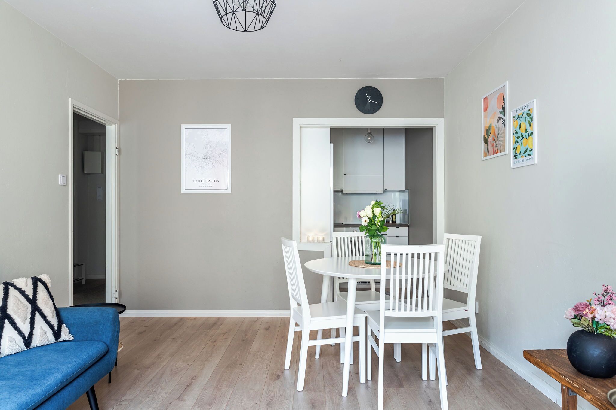A modern dining area with a white table and chairs, a clock on the wall, and a kitchen visible through an open door.