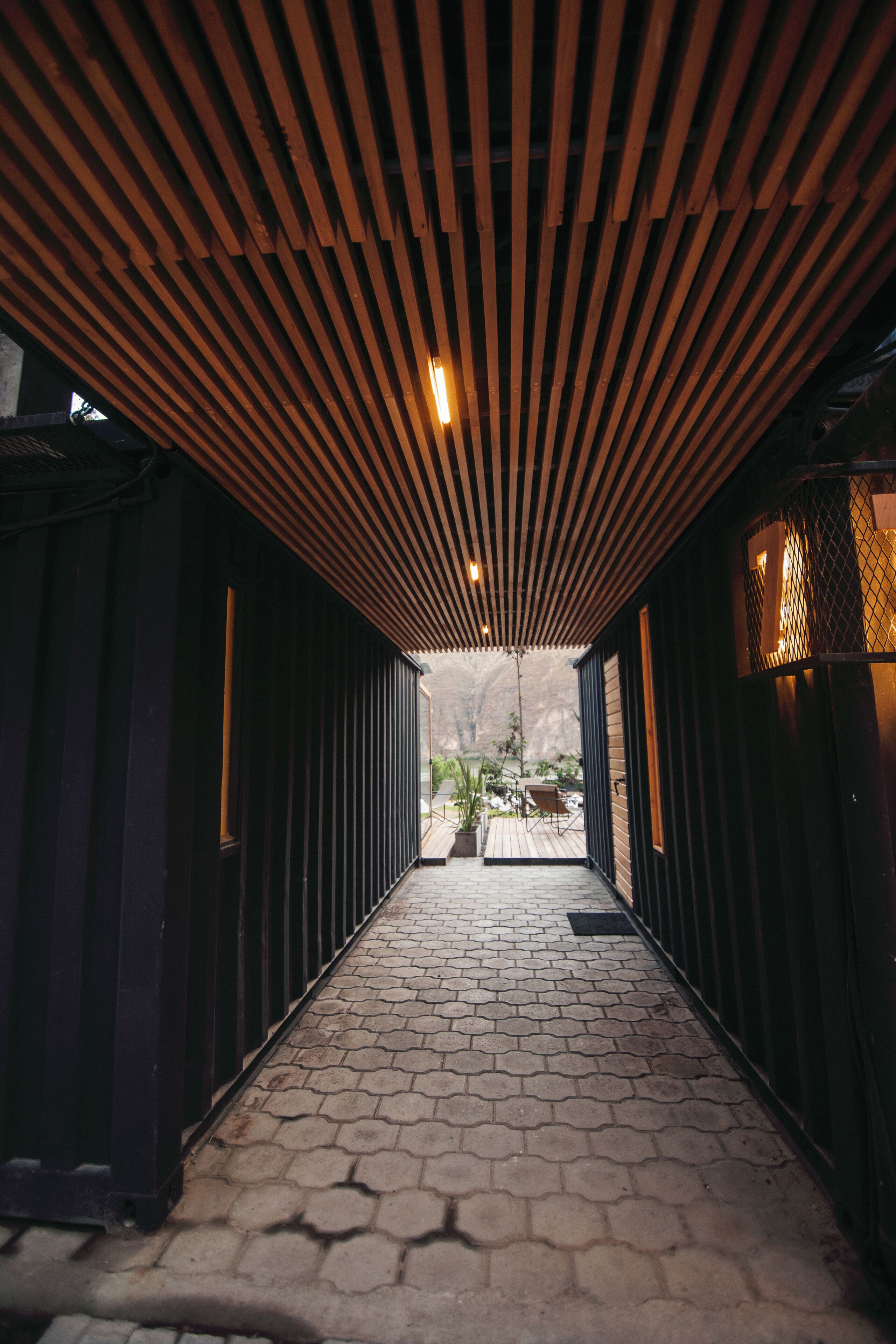 A corridor with a wooden slatted ceiling and hexagonal paving leading to a view of a mountainous landscape.