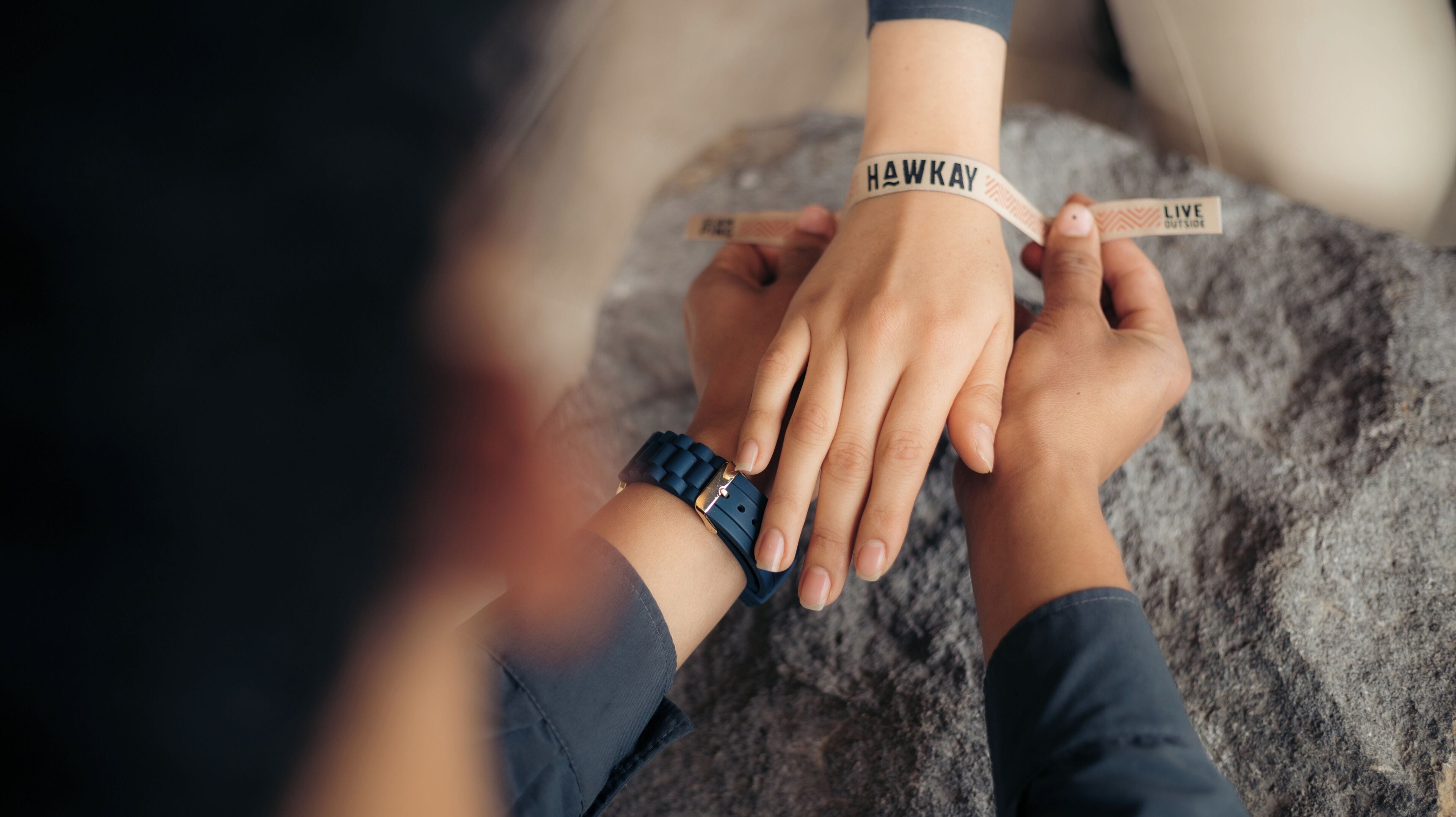 Two hands holding a wristband with the text "HAWKAY" and "LIVE OUTSIDE".