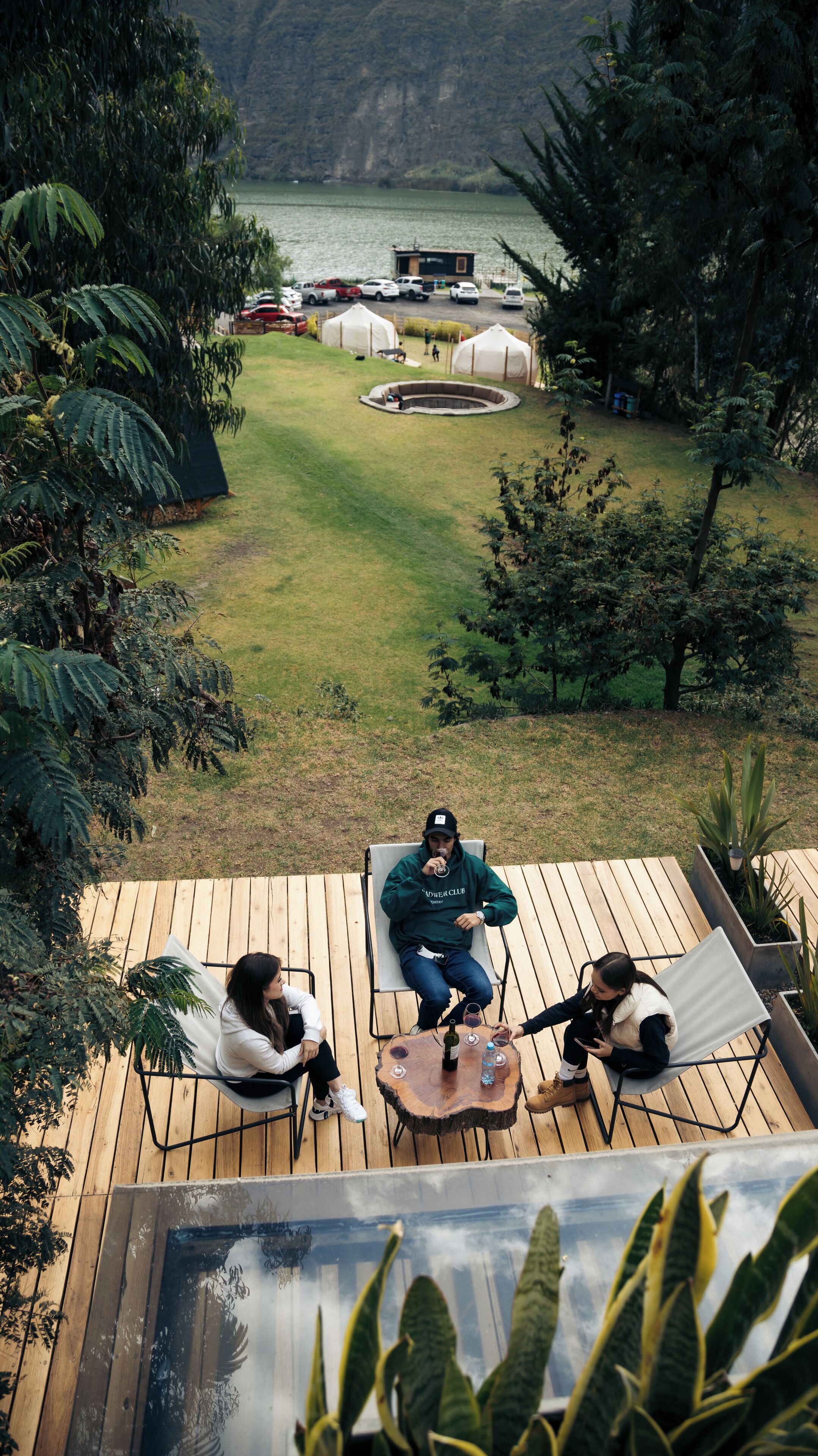 Um deck com três cadeiras, uma mesa de madeira com uma garrafa e copos, e vista para um lago com tendas ao fundo.