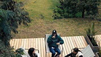 Um deck com três cadeiras, uma mesa de madeira com uma garrafa e copos, e vista para um lago com tendas ao fundo.