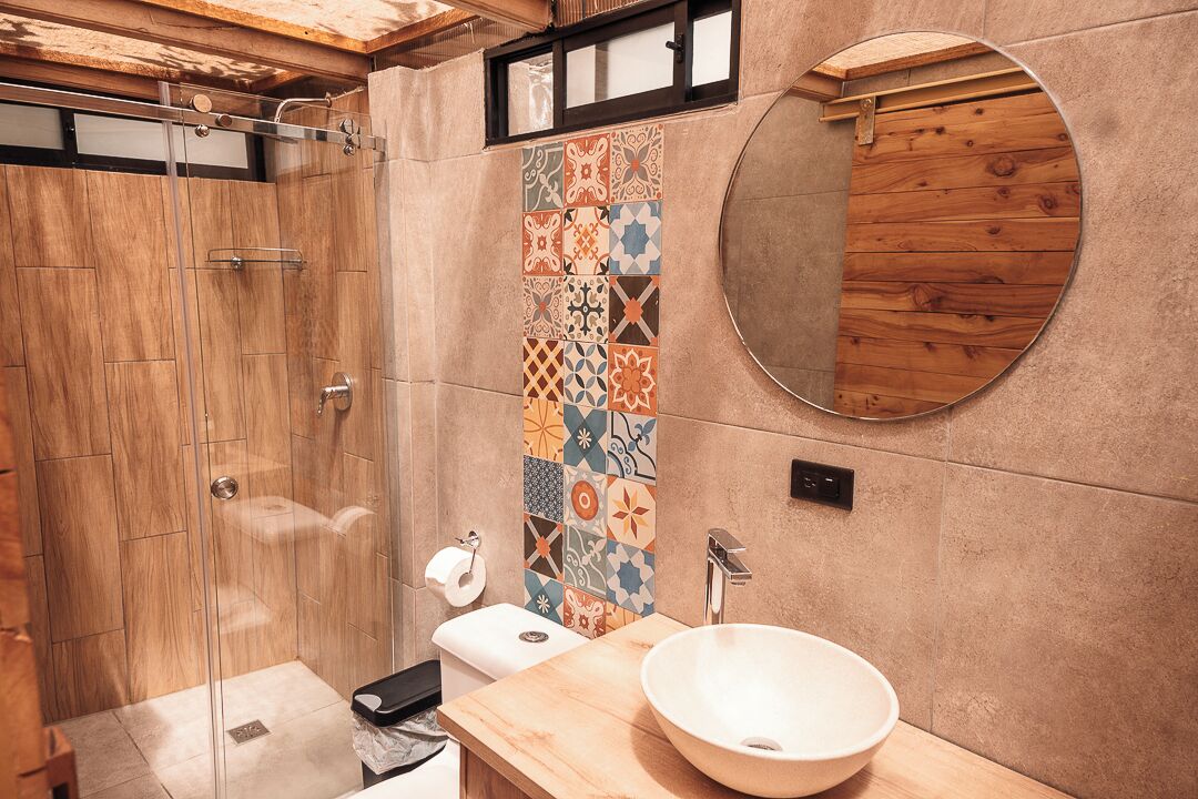 A modern bathroom with a wooden shower enclosure, a tiled wall with a colorful pattern, a round mirror, and a wooden vanity with a sink.