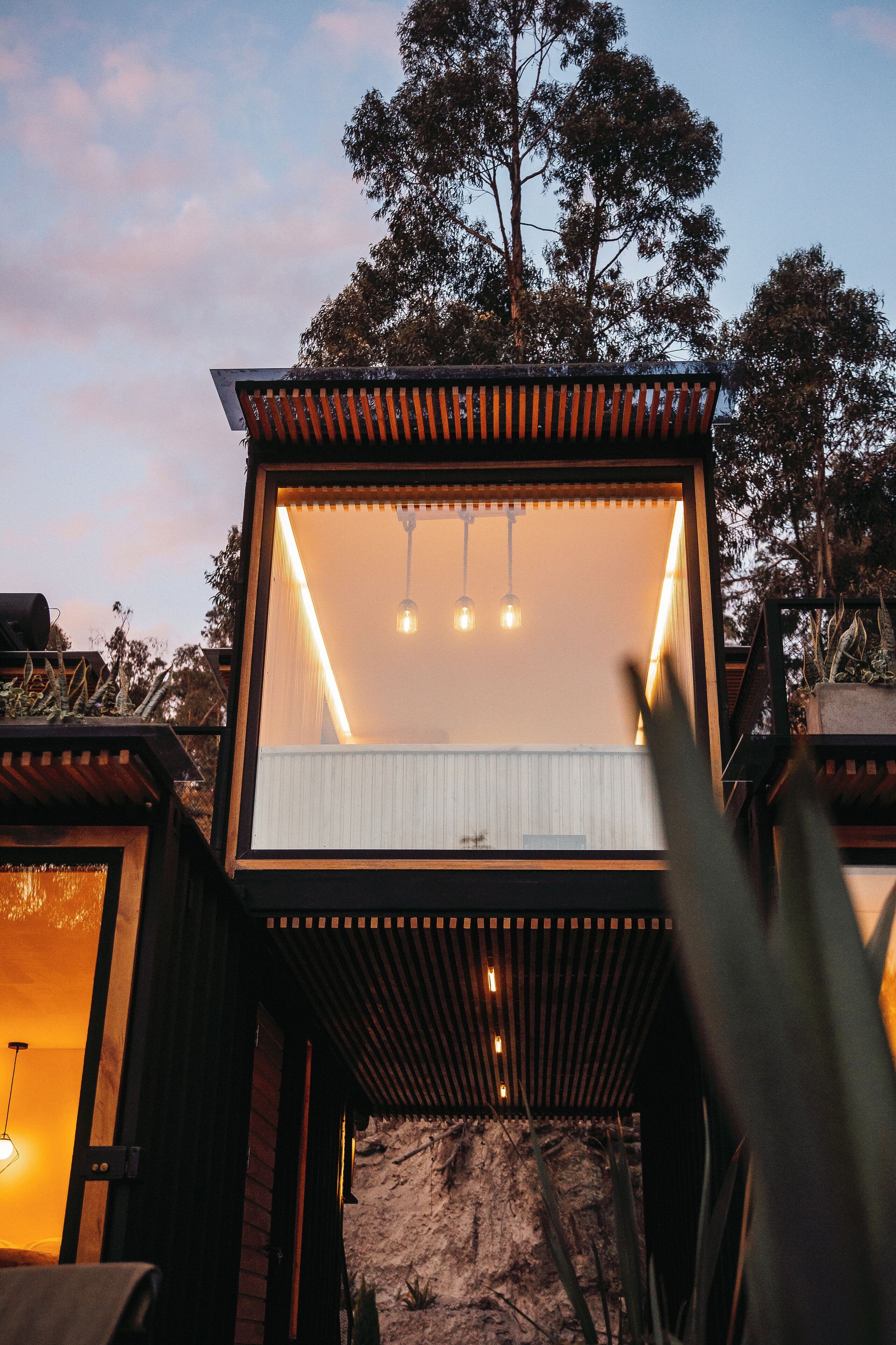 Una estructura de madera moderna y elevada con cerramientos de vidrio, iluminación interior y vista de los árboles circundantes.