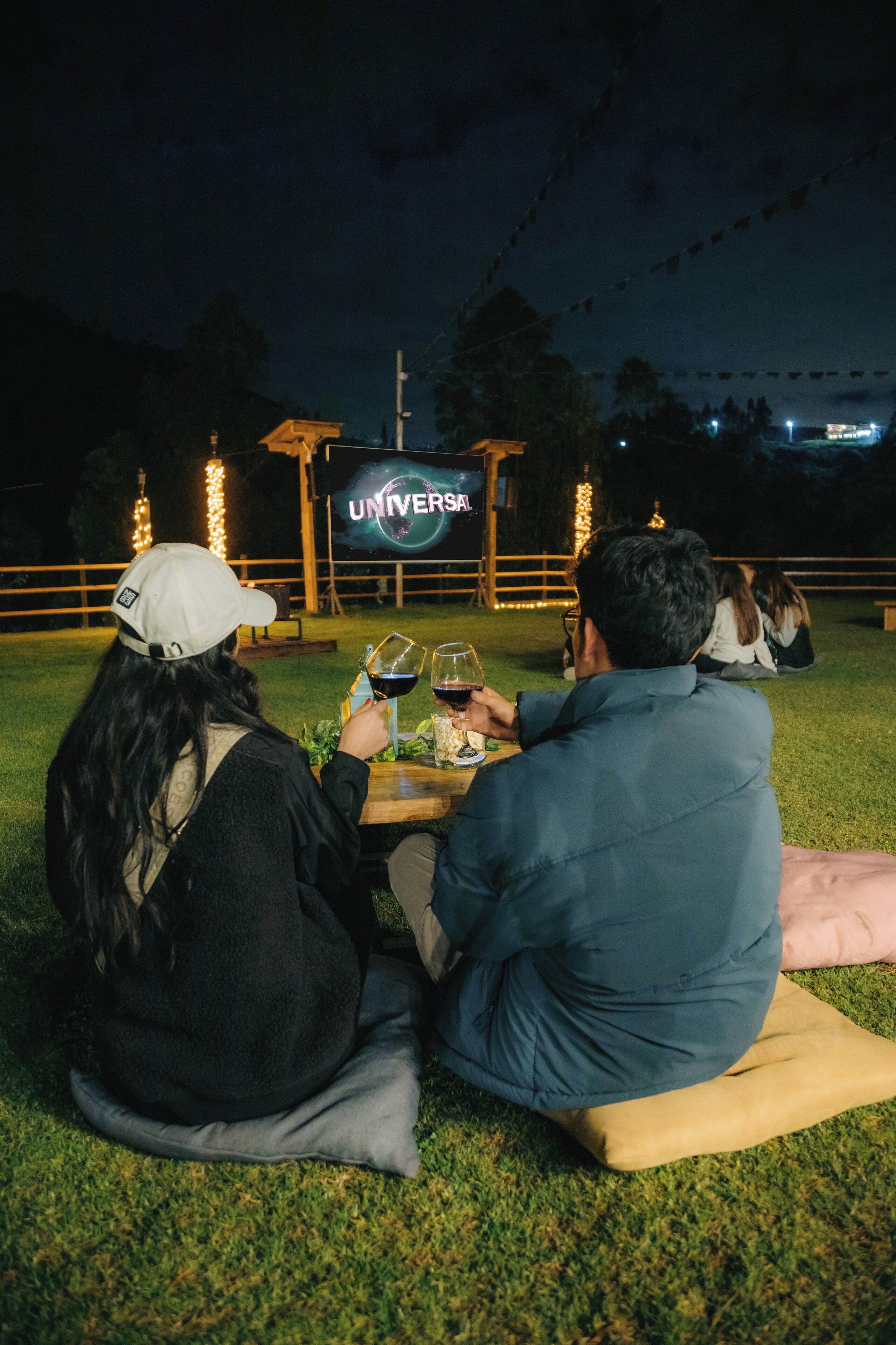Two people sitting on the grass at a table with wine glasses, under a sign that reads "UNIVERSAL".
