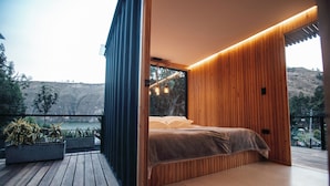 A modern bedroom with a wooden slatted wall, a bed with a beige blanket, and a view of a hillside. - Hawkay Glamping (Panzaleo)