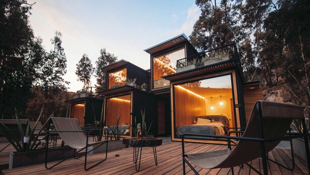 A modern outdoor living area with wooden flooring, seating, and a view of a bedroom with a bed and a nightstand. - Hawkay Glamping (Panzaleo)
