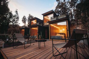 Un'area relax esterna moderna con pavimento in legno, posti a sedere e vista su una camera da letto con un letto e un comodino.