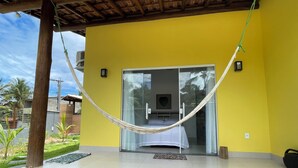 Un bâtiment jaune avec un hamac devant une porte-fenêtre coulissante qui donne sur une pièce où se trouve un lit.