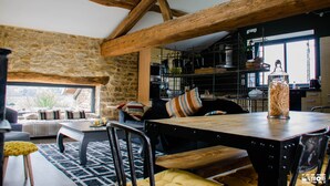 A rustic interior with exposed wooden beams, a stone wall, a dining area with a wooden table, and a kitchen with shelves and appliances. - Gîte de La Pyranvôle (Lacrost)