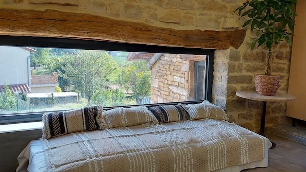 A cozy bedroom with a large window, a bed with patterned bedding, a potted plant, and a modern kitchen in the background. - Gîte de La Pyranvôle (Lacrost)