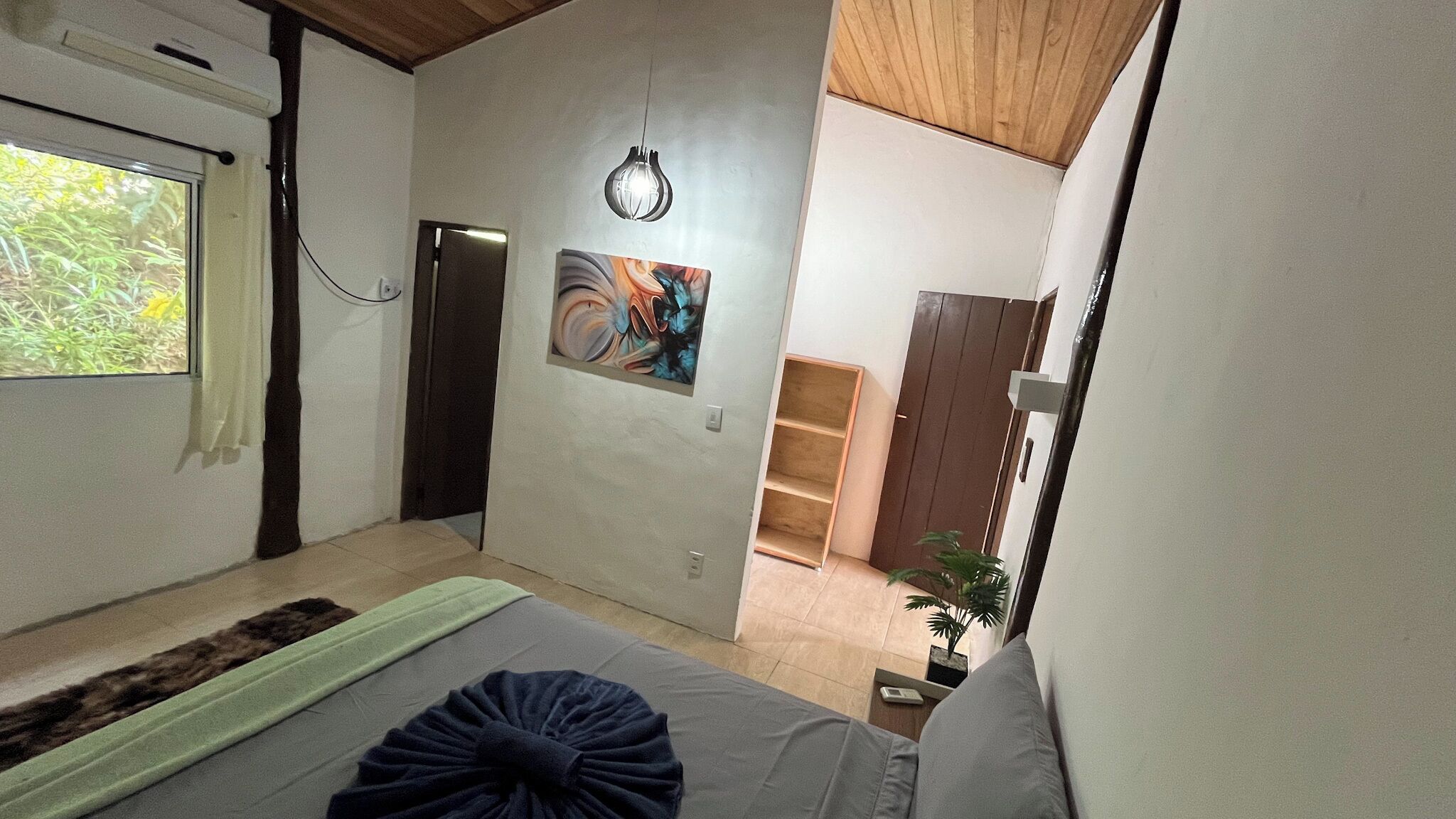 A bedroom with a bed, a window, a door, a wall-mounted light, and a painting.