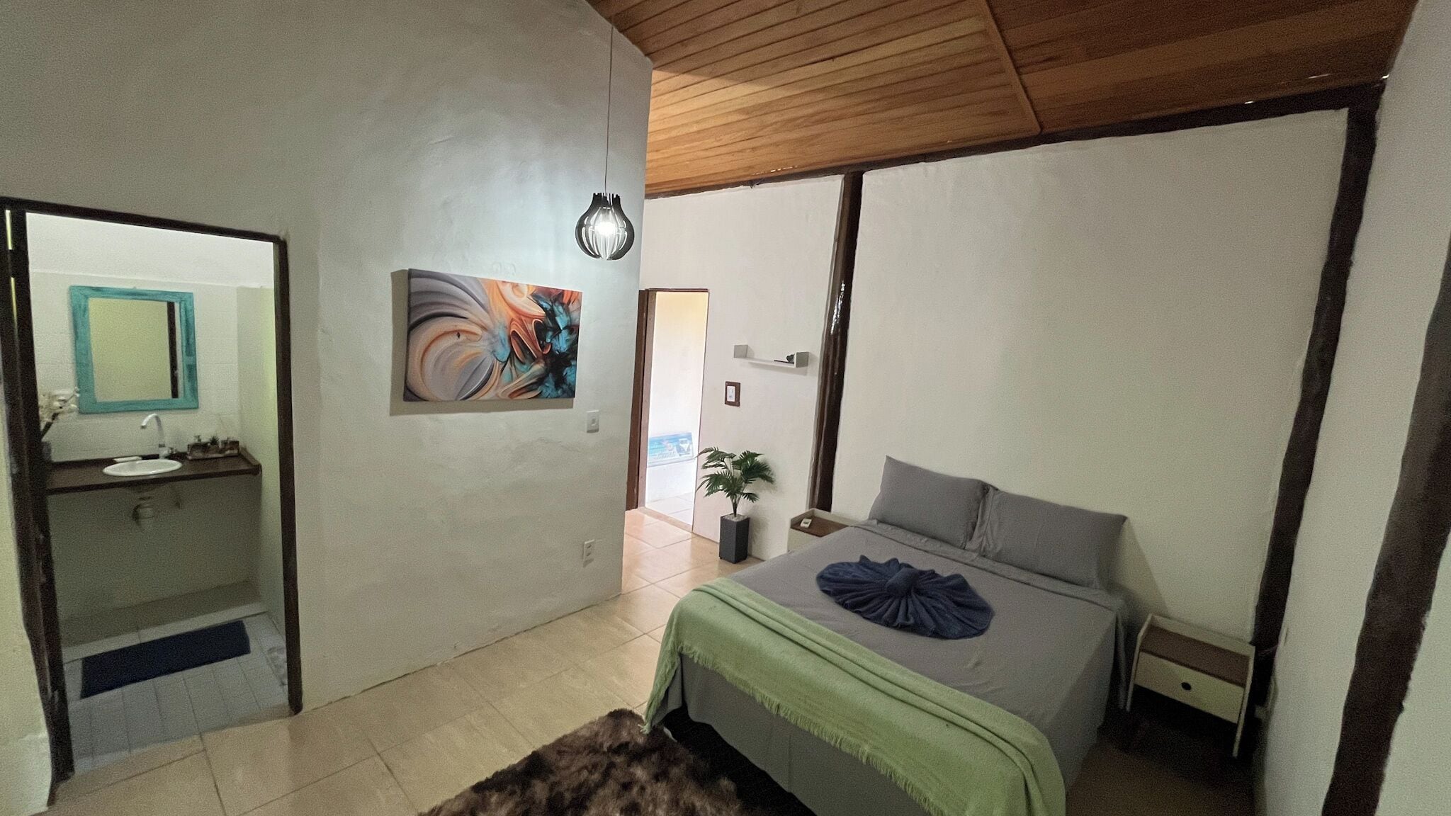 A bedroom with a bed, a bedside table, a nightstand, a mirror, a sink, and a potted plant.