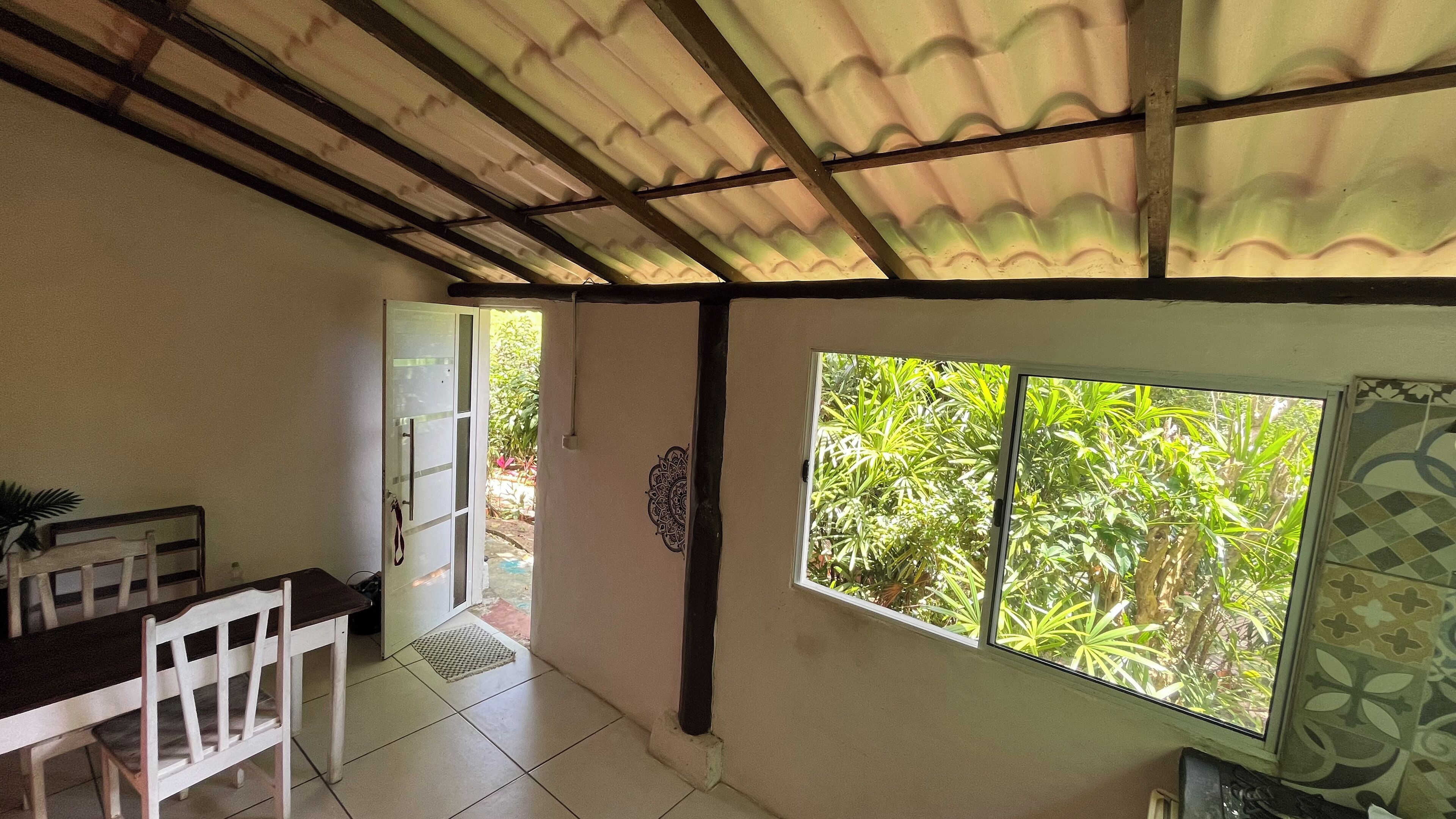 A room with a tiled floor, a dining table with chairs, a window with a view of greenery, and a door leading outside.