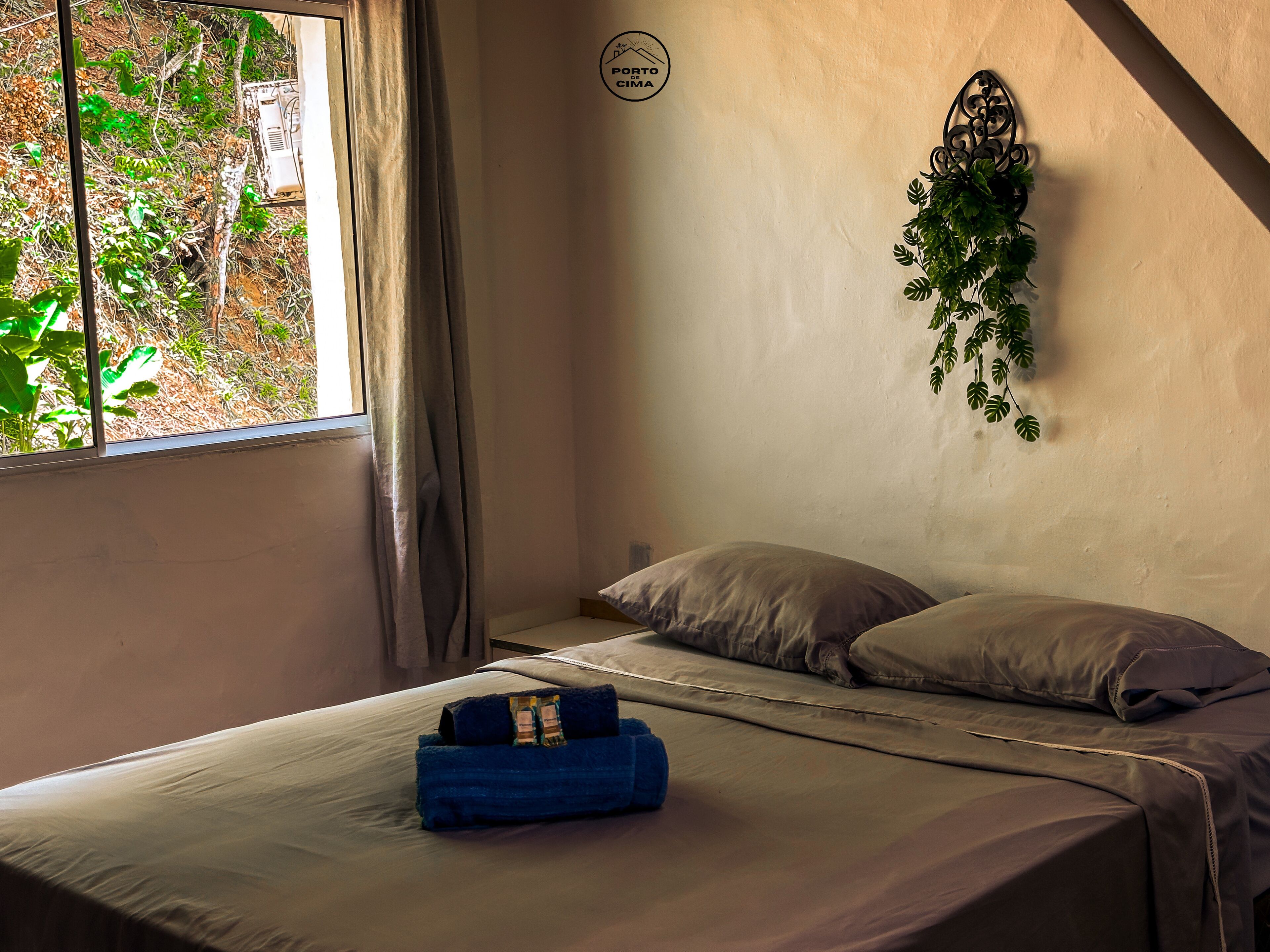Una cama individual con una toalla azul y un almohadón, una ventana con cortinas y una planta sujeta a la pared.