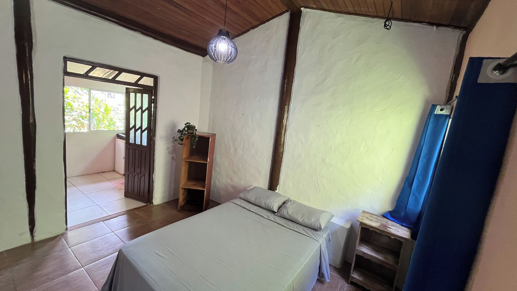 A bedroom with a bed, a wooden shelf, a hanging light fixture, and a door leading to another room.
