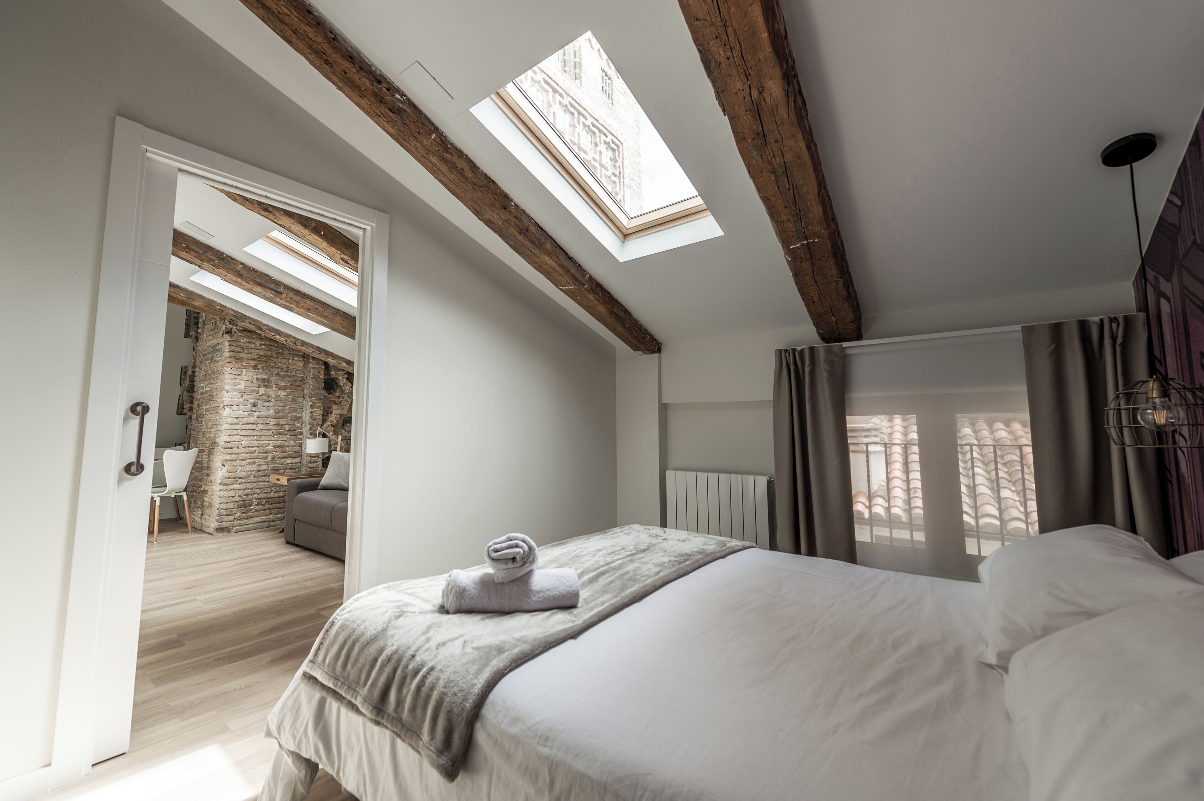 Un dormitorio con tragaluz, una cama con una toalla enrollada, un radiador y vistas a un muro de ladrillo y un balcón.