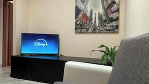 Een woonkamer met een flatscreen-tv waarop het Disney+-logo te zien is, een groot stadslandschap aan de muur en een potplant.