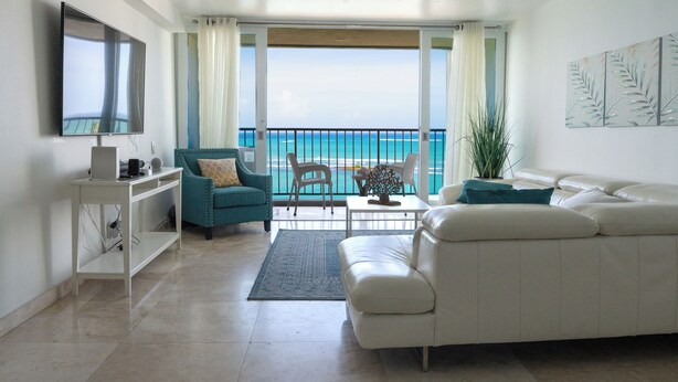Een woonkamer met een witte bank, blauwgroene fauteuils, een salontafel en een tv op een witte kast. Er is een balkon met uitzicht op de oceaan.