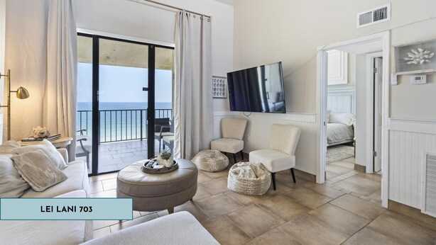 A modern living room with a sofa, ottoman, and chairs, a television, and a view of the sea through large windows. - Lei Lani 703 (Orange Beach)