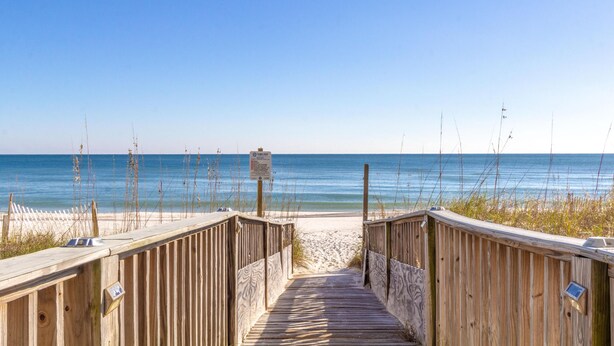 Ein hölzerner Steg führt zu einem Sandstrand, über dem ein klarer, blauer Himmel erstrahlt.
