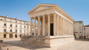 Un bâtiment de style classique, doté d’une entrée centrale flanquée de colonnes, entouré d’une place pavée et bordé d’autres bâtiments.