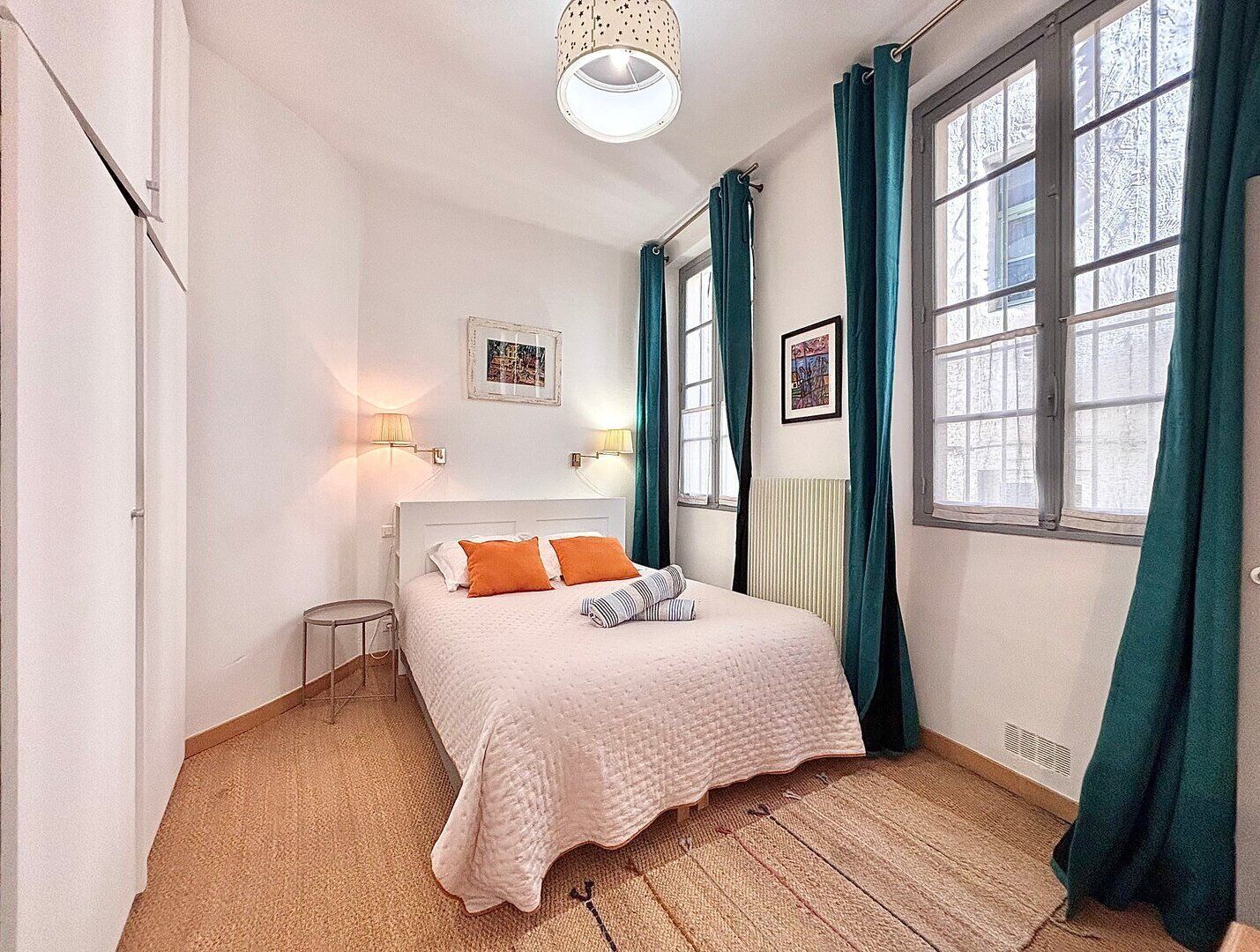 Un dormitorio con una cama, dos lámparas de pared, un ventanal con cortinas y un radiador.