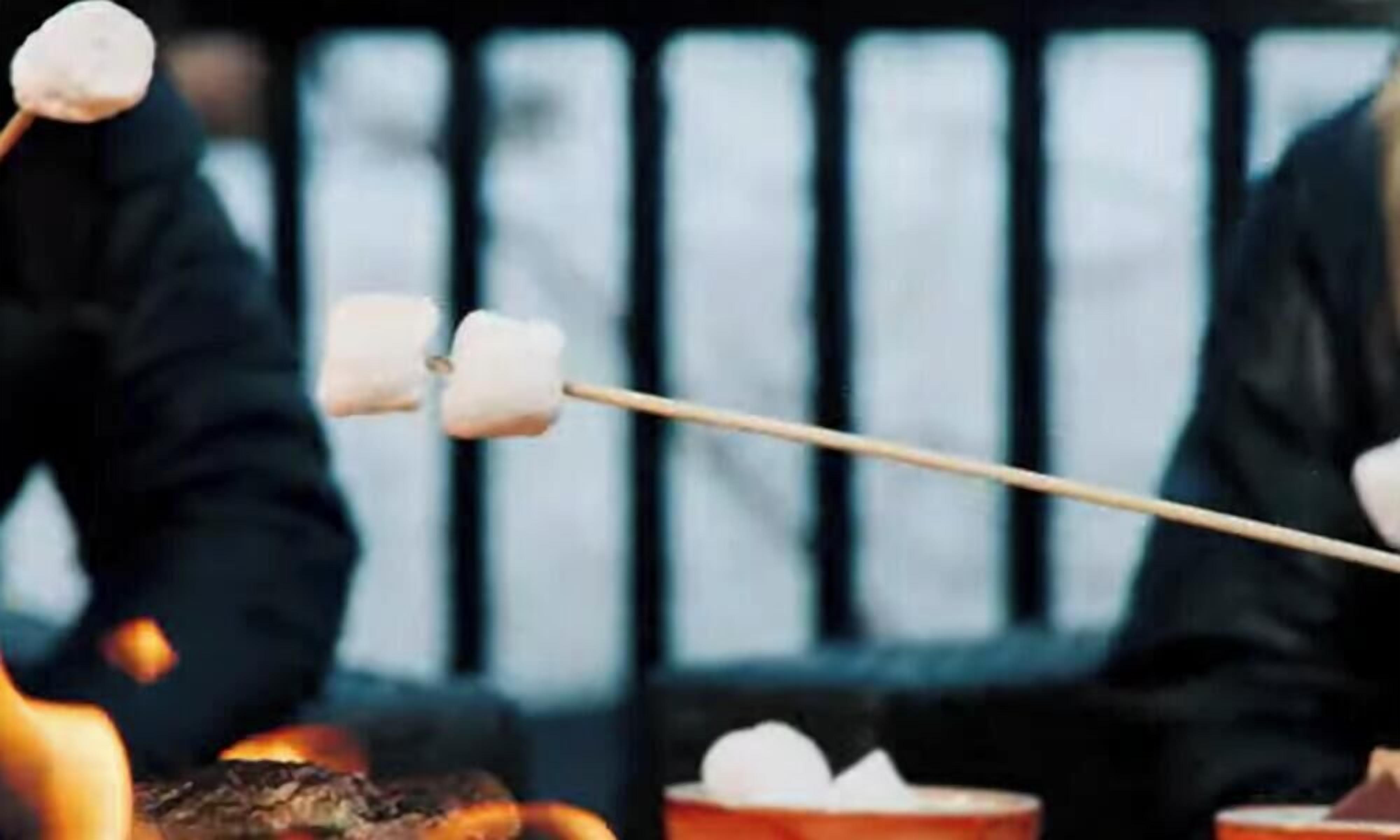 Nahaufnahme von zwei Spießen mit Marshmallows, die am Feuer geröstet werden.